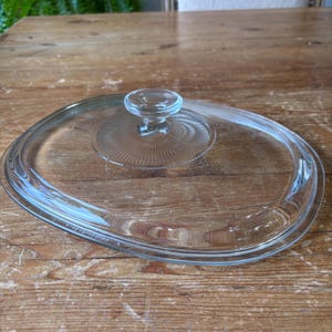 May include: A clear, oval-shaped glass lid with a circular handle. The lid has a ridged design around the handle and a smooth, curved edge. The lid is designed for cookware.