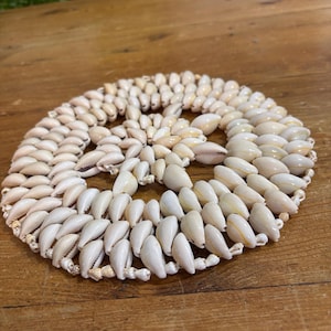 May include: Circular coaster made of numerous small, cream-colored seashells. The shells are arranged in a spiral pattern, creating a decorative and natural aesthetic. The coaster is designed to protect surfaces from heat and moisture.