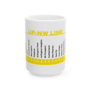 May include: A white ceramic mug featuring a yellow horizontal band with the text UP-NW LINE in bold yellow letters. Below the text, a list of train stations including Barrington, Palatine, and Des Plaines is printed in black, with a yellow line.