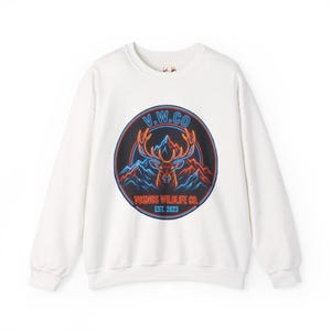 Puede incluir: Sudadera blanca de cuello redondo con un diseño gráfico circular. El diseño presenta una cabeza de ciervo, montañas y el texto "V.W.CO YOSHOS WILDLIFE CO. EST. 2023". El diseño utiliza colores azul, rojo y naranja.