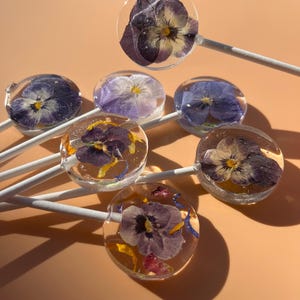May include: A collection of handcrafted lollipops, each featuring a vibrant, pressed flower encased in clear candy. The flowers display shades of purple, yellow, and white, set against a light peach background. The lollipops are on white sticks.