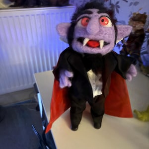 May include: A needle-felted vampire doll with purple skin, red eyes, and white fangs. The doll wears a black suit jacket with a white shirt underneath and a long cape with a vibrant red interior lining. The figure has dark hair and a pointed goatee.