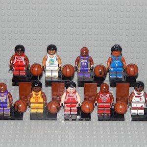 May include: A collection of Lego basketball player figurines, each holding a brown basketball. The figures wear jerseys in various team colors, including red, yellow, blue, and purple. The background is a gray Lego baseplate.