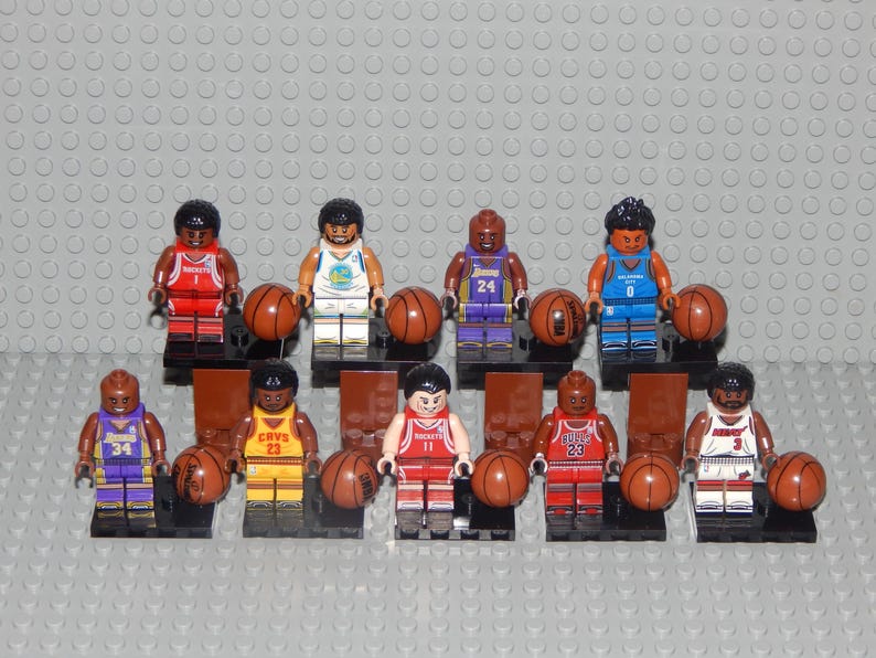 May include: A collection of Lego basketball player figurines, each holding a brown basketball. The figures wear jerseys in various team colors, including red, yellow, blue, and purple. The background is a gray Lego baseplate.
