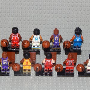 May include: A collection of Lego basketball player figurines, each holding a brown basketball. The figurines are wearing jerseys with different numbers and team colors. The background is a gray Lego baseplate.