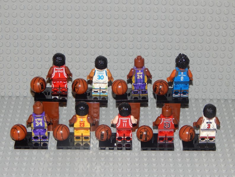 May include: A collection of Lego basketball player figurines, each holding a brown basketball. The figurines are wearing jerseys with different numbers and team colors. The background is a gray Lego baseplate.