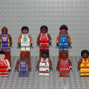 May include: A collection of ten Lego minifigures, each representing a basketball player. The figures wear jerseys in various team colors, including purple, red, blue, and yellow. Each figure has a unique number on their jersey.