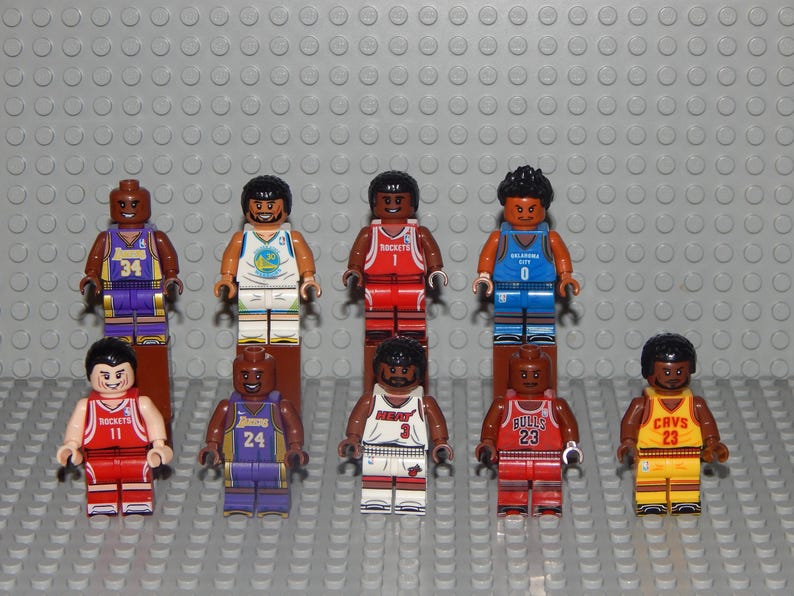 May include: A collection of ten Lego minifigures, each representing a basketball player. The figures wear jerseys in various team colors, including purple, red, blue, and yellow. Each figure has a unique number on their jersey.