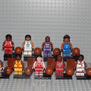 May include: A collection of Lego basketball player minifigures, each holding a brown basketball. The figures wear team jerseys in various colors, including red, yellow, blue, and purple. The background is a gray surface.