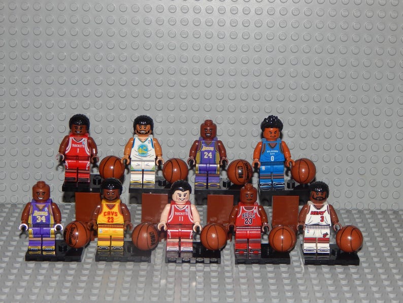 May include: A collection of Lego basketball player minifigures, each holding a brown basketball. The figures wear team jerseys in various colors, including red, yellow, blue, and purple. The background is a gray surface.