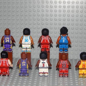 May include: A collection of miniature basketball player figures in various team colors. Each figure is wearing a jersey with a player's name and number. The figures are arranged on a gray surface with a textured background.