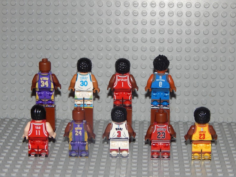 May include: A collection of miniature basketball player figures in various team colors. Each figure is wearing a jersey with a player's name and number. The figures are arranged on a gray surface with a textured background.