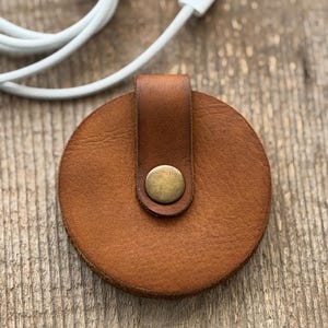 May include: A round, brown leather cable organizer with a brass snap closure. The organizer has a loop for securing the cable and is shown with a white cable.
