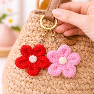 Könnte beinhalten: Zwei Schlüsselanhänger mit gehäkelten Blumen, einer rot und einer rosa, mit weißen Zentren, hängen an goldfarbenen Schlüsselringen. Die Schlüsselanhänger sind an einer gewebten Tasche mit braunem Ledergriff befestigt.