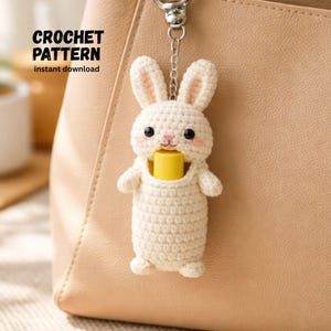 May include: A cream-colored crochet bunny keychain with a yellow object in its mouth, attached to a silver keyring. The bunny has pink accents on its ears and cheeks. Text on the left reads "CROCHET PATTERN instant download".