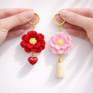 May include: Two handmade crochet flower keychains. One is red with a yellow center and a heart charm. The other is pink with a yellow center and a cream tassel. Both have gold-colored key rings.
