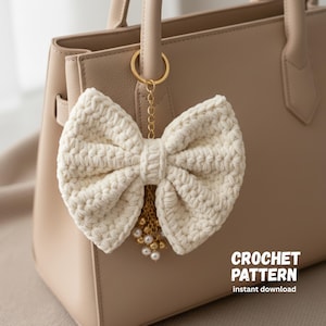 May include: A cream-colored, crocheted bow-shaped keychain with a gold chain and pearl accents, hanging from a beige handbag. The keychain is attached to a gold ring. Text on the image reads "CROCHET PATTERN instant download."