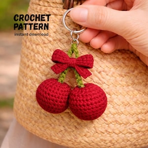 Puede incluir: Llavero de ganchillo con dos cerezas rojas y un lazo rojo, unidos por tallos verdes. Se adjunta a un anillo de metal plateado. El texto "CROCHET PATTERN instant download" es visible.