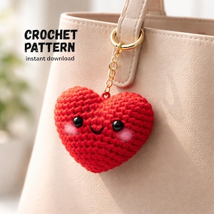 May include: A red crocheted heart-shaped keyring with a smiling face and pink cheeks, suspended from a gold chain. The text "CROCHET PATTERN instant download" is in the upper left corner. The keyring is attached to a beige handbag.