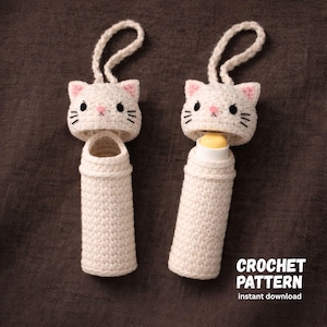 Pode incluir: Duas capas de bálsamo labial em crochê branco em forma de gato com detalhes em rosa. Cada capa tem uma alça para pendurar e um design de rosto de gato. Uma capa está aberta e a outra contém um bálsamo labial. O texto diz "CROCHET PATTERN instant download".
