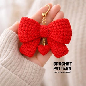 Puede incluir: Un llavero de lazo rojo tejido a ganchillo, con un cierre dorado y un pequeño dije de corazón. El lazo es detallado y texturizado, con un colgante de corazón a juego. El texto "CROCHET PATTERN instant download" es visible.
