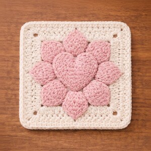 May include: A square, off-white crochet piece with a central pink heart shape surrounded by eight pink petal shapes. The piece is set against a wooden background, showcasing the handmade texture and design.