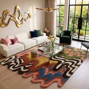 May include: A large area rug featuring a multi-colored, wavy, abstract pattern with flowing lines in shades of brown, green, blue, pink, and cream. The rug has an irregular, organic shape with curved edges that extend across the light wood floor.