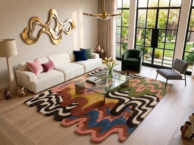 May include: A large area rug featuring a multi-colored, wavy, abstract pattern with flowing lines in shades of brown, green, blue, pink, and cream. The rug has an irregular, organic shape with curved edges that extend across the light wood floor.