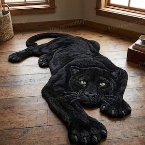 Könnte beinhalten: Ein schwarzer Panther-Teppich mit grünen Augen, der auf einem Holzboden liegt. Der Teppich besteht aus einem weichen, strukturierten Material und weist detaillierte Pfoten, einen Schwanz und ein Gesicht auf. TheHeavenRugs ist auf dem Teppich aufgedruckt.