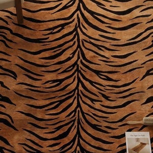 Op de afbeelding: Een bruin en zwart tapijt met tijgerstrepenpatroon. Het tapijt heeft een herhalend patroon van zwarte strepen op een bruine achtergrond. Het tapijt is rechthoekig en lijkt van een zacht materiaal te zijn. Een klein boek met de titel "The Tiger Tie Path" ligt op het tapijt.