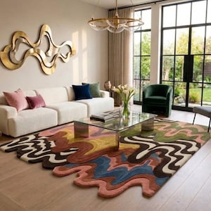 May include: A large, irregularly shaped area rug featuring a multi-colored, wavy, abstract pattern in shades of pink, blue, yellow, brown, and green. The rug has a textured, tufted surface and sits on a light-colored wooden floor in a living room.