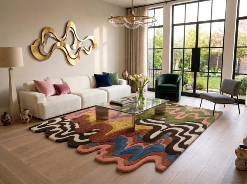 May include: A large, irregularly shaped area rug featuring a multi-colored, wavy, abstract pattern in shades of pink, blue, yellow, brown, and green. The rug has a textured, tufted surface and sits on a light-colored wooden floor in a living room.