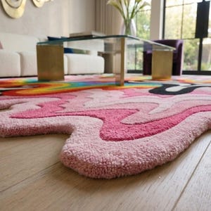 May include: A tufted area rug with an irregular, organic shape featuring a vibrant, multi-colored design. The surface displays wavy, flowing patterns in shades of light pink, hot pink, black, yellow, orange, and blue, creating a bold, liquid-like effect.