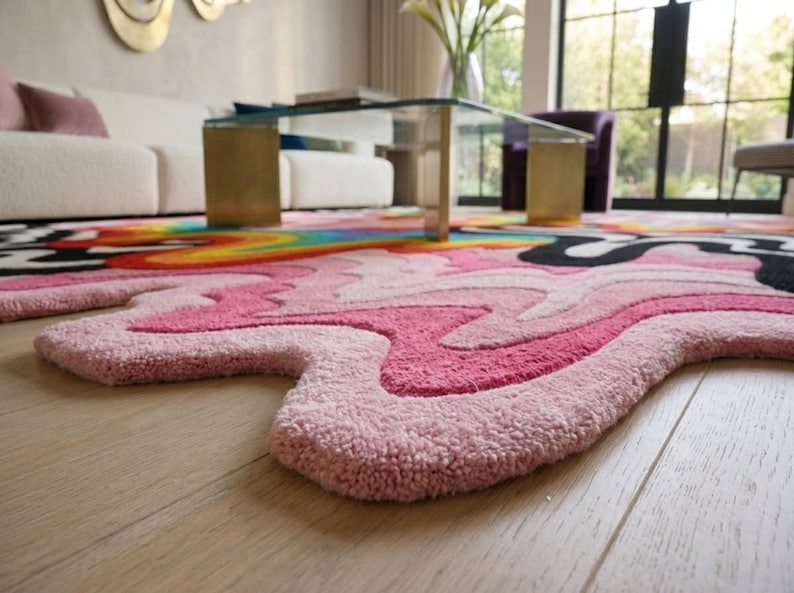 May include: A tufted area rug with an irregular, organic shape featuring a vibrant, multi-colored design. The surface displays wavy, flowing patterns in shades of light pink, hot pink, black, yellow, orange, and blue, creating a bold, liquid-like effect.