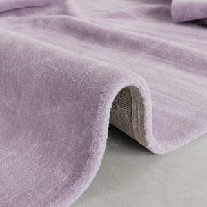 May include: A close-up of a soft, lavender-colored rug with a rolled edge. The plush texture of the rug is visible, with a neutral-colored backing. The rug is likely made of a soft material, suitable for a living room or bedroom.