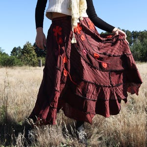 Op de afbeelding: Een vrouw draagt een wit gebreid vest over een zwart shirt met lange mouwen, een bordeauxrode rok met bloemenapplicaties en een crèmekleurige sjaal. De rok heeft een gelaagd, gerimpeld ontwerp. De achtergrond is een veld met hoog gras en een heldere blauwe lucht.