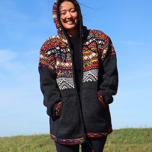 Op de afbeelding: Een donkergrijze gebreide jas met capuchon en een kleurrijk geometrisch patroon op de capuchon en borst. De jas heeft een volledige ritssluiting, twee voorzakken en een trekkoord aan de onderkant. Het patroon bevat oranje, rode, witte en blauwe accenten.