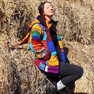 Op de afbeelding: Een kleurrijke patchworkjas met capuchon, met een levendige mix van regenboogkleuren, blauw, rood en geel. De jas heeft een zwarte kraag en wordt gedragen met zwarte leggings en laarzen. De capuchon van de jas heeft een decoratieve kwast.