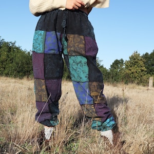 Könnte beinhalten: Patchworkhose mit schwarzem Bund und Kordelzug. Die Hose hat eine Vielzahl von bunten Quadraten in Blau-, Lila-, Türkis- und Goldtönen. Die Hose wird im Freien in einem Feld mit hohem Gras getragen.