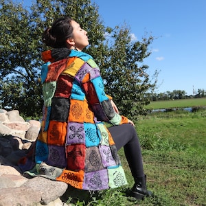 Könnte beinhalten: Ein Patchwork-Mantel in leuchtenden Farben, darunter Orange, Blau und Lila, mit schwarzen dekorativen Mustern. Der Mantel hat eine Kapuze und wird im Freien getragen. Der Mantel besteht aus mehreren zusammengenähten Stoffquadraten.
