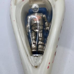 May include: A vintage toy rocket ship in white with red accents. A silver astronaut figure is visible inside a clear plastic bubble. The rocket has red paint splatters and a fin on top.