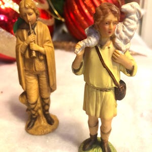 Può includere: Due statuine raffiguranti personaggi biblici. Una figura suona il flauto, l'altra porta un agnello sulle spalle. Entrambe le figure indossano abiti color beige e poggiano su basi verdi. Ornamenti natalizi sullo sfondo.