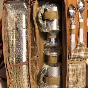 May include: Open brown picnic set with stainless steel cups, utensils, and a plaid napkin. The set includes two spoons, two cups, and a container for other items. The set is in a brown zippered case.