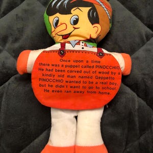 May include: A Pinocchio doll with a smiling face, orange overalls, and white legs. The doll has a printed story on its chest. The text reads, "Once upon a time there was a puppet called PINOCCHIO."