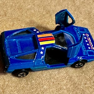 May include: A blue die-cast toy car with the doors open. The car has white star decals and a multi-colored stripe on the roof. The text "DOMO-Q" is visible on the side of the car.