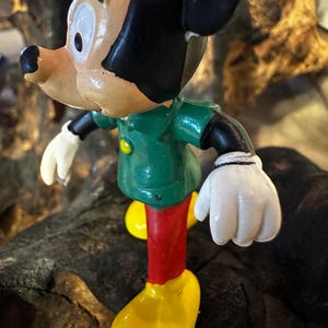 May include: A vintage figurine of a cartoon character, featuring a green jacket, red pants, and yellow shoes. The character has black hair, a tan face, and white gloves. The figurine is posed on a dark, textured surface.