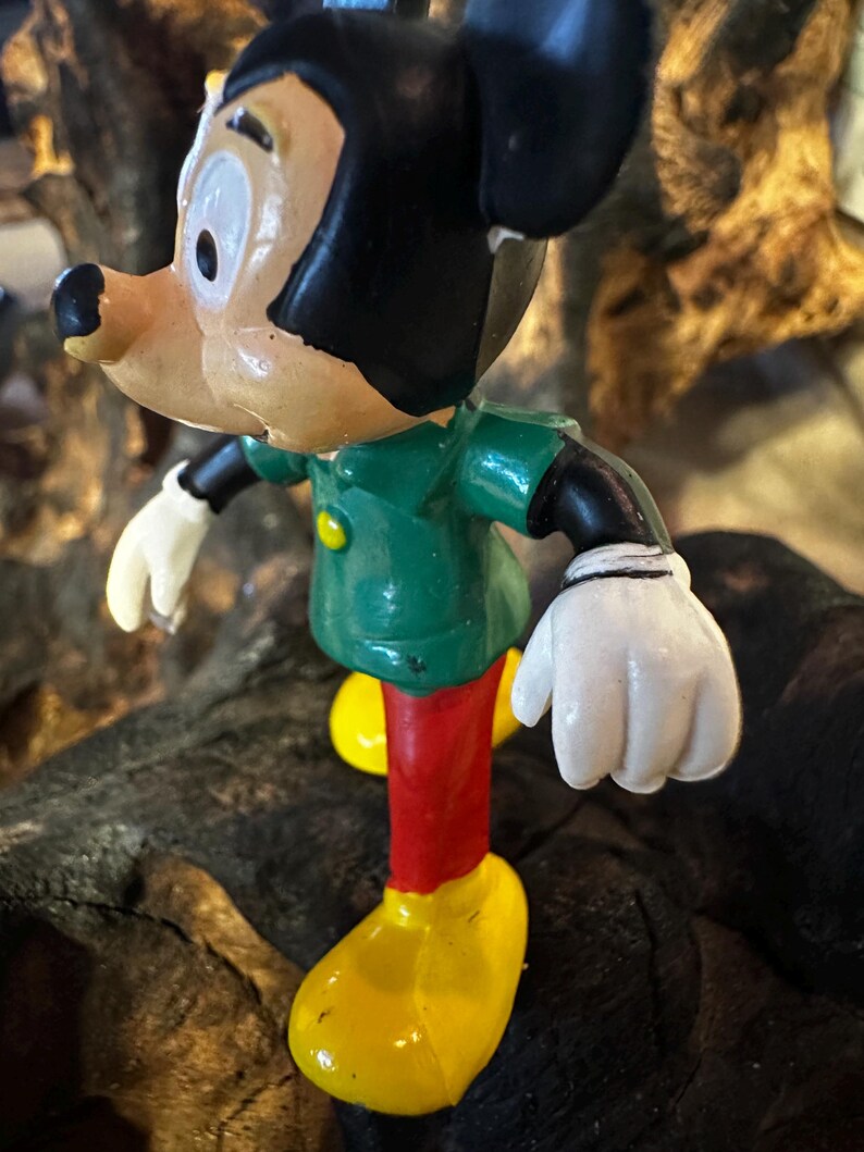 May include: A vintage figurine of a cartoon character, featuring a green jacket, red pants, and yellow shoes. The character has black hair, a tan face, and white gloves. The figurine is posed on a dark, textured surface.