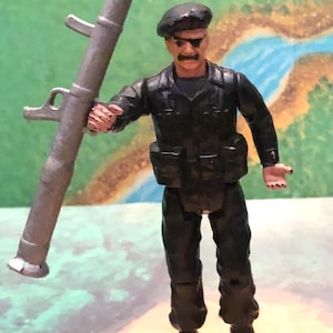 May include: A toy soldier figure in a black uniform and beret, holding a gray rocket launcher. The figure has a mustache and sunglasses. The background features a painted landscape with green and blue colors.