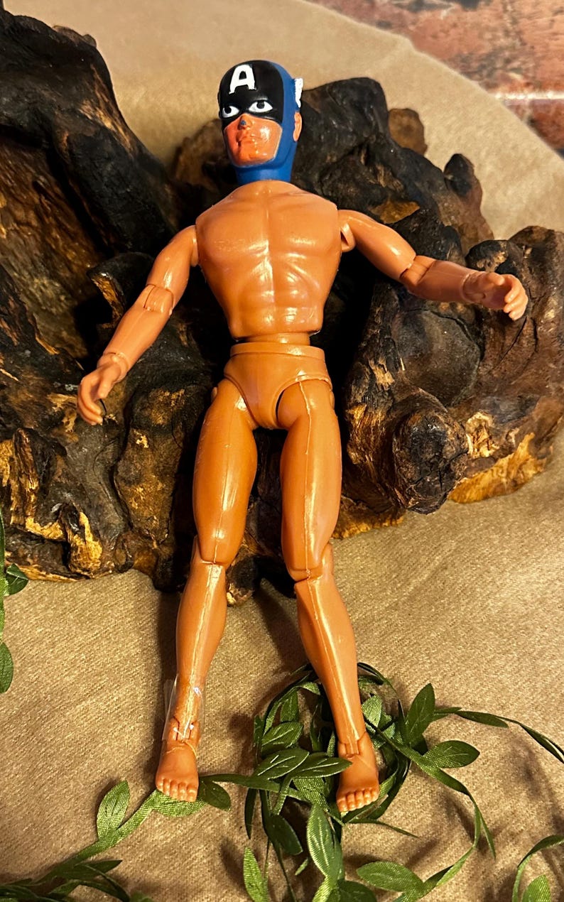 May include: A vintage action figure of a superhero, featuring a blue mask with a white "A" on a black background. The figure is tan with articulated joints and stands in a dynamic pose. The figure is approximately 12 inches tall.