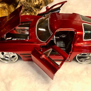 May include: A detailed, red model car with open doors and hood, showcasing the engine and interior. The car has chrome wheels and a glossy finish, capturing the classic car aesthetic.